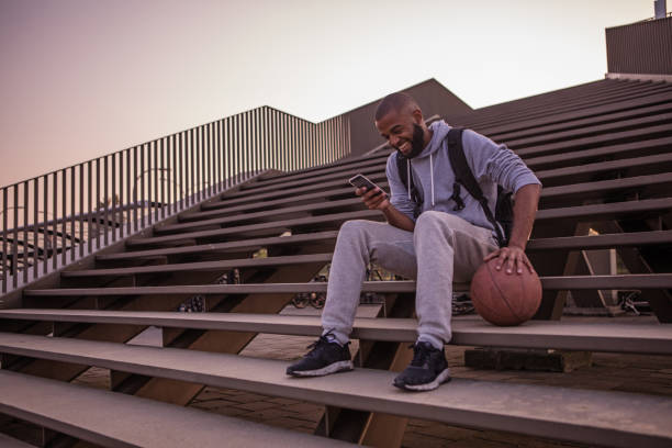 un giovane studente di basket maschile che gioca a basket usando il suo smartphone mentre è seduto su alcuni gradini della città - streetball basketball sport men foto e immagini stock