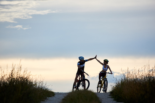 Sporty woman and man wearing sportswear and helmets, riding bicycles, having fun outside. Silhouettes of sportsmen highing five and posing on road in sunset time. Non urban scene.