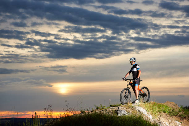 istock Cyclist with bicycle rests on cliff top in mountains enjoying sun at sunset under an unusual sky 961945184