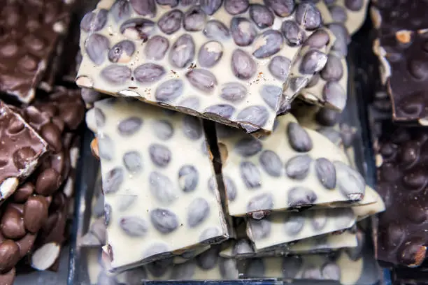 Macro closeup of pile of many white chocolate pieces chunks bars with peanuts, almonds on tray display in candy store chocolatier shop