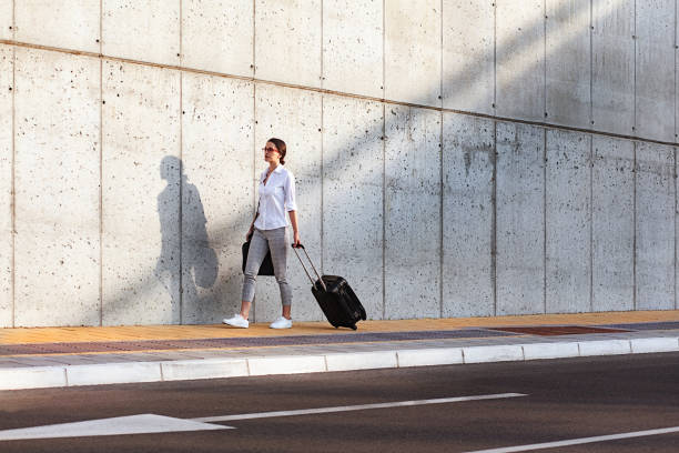 istock Young woman walking on a sidewalk beside the concrete wall and pulling a small wheeled luggage 961963236