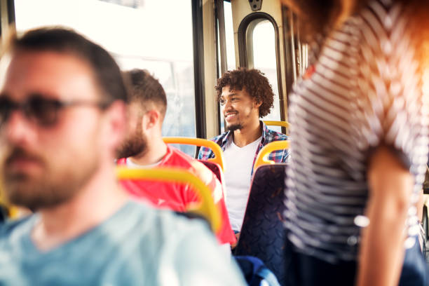 istock Young cheerful handsome man is sitting on a bus looking through a window and smiling while being surrounded by other passengers. 961971580