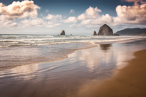 Sunset looms over Haystack Rock in Cannon Beach along the Oregon Coast, Pacific Northwest Landmark and tourist destination. Light falls onto crashing waves of incoming Tide. Reflections of cloudscape onto receding surf and sand.