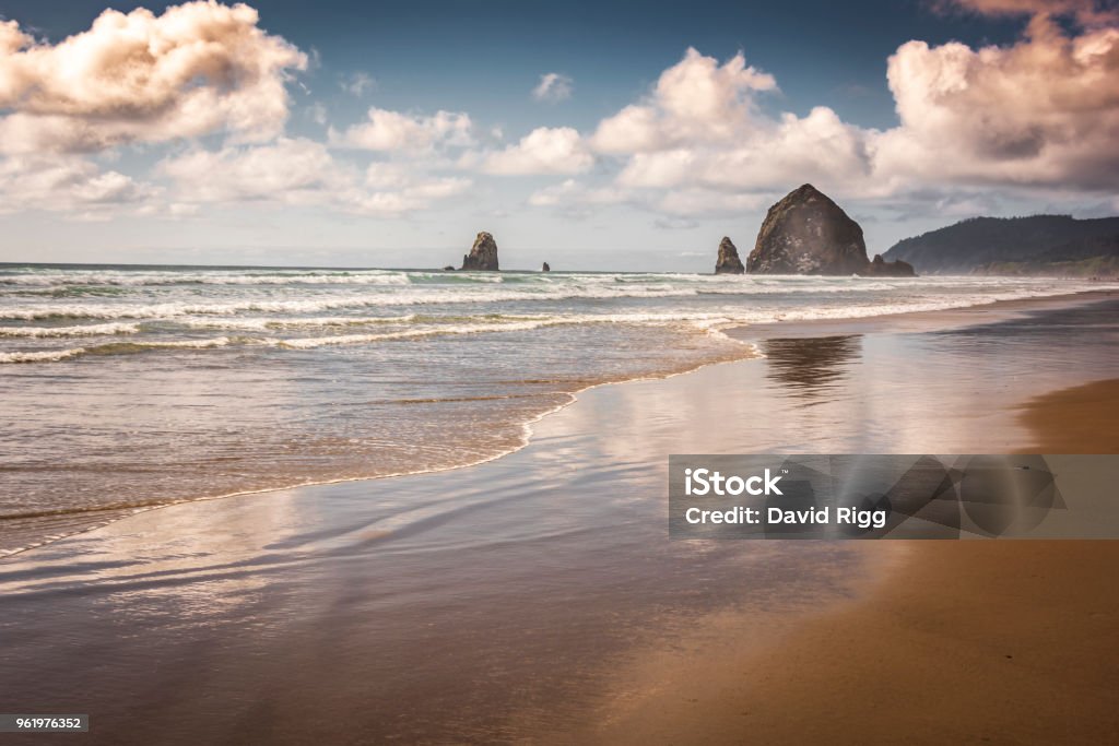 Canon Beach Oregon Haystack Rock Reflections Clouds Tide Surf Beach Waves Sunset Sunset looms over Haystack Rock in Cannon Beach along the Oregon Coast, Pacific Northwest Landmark and tourist destination. Light falls onto crashing waves of incoming Tide. Reflections of cloudscape onto receding surf and sand. Cannon Beach Stock Photo