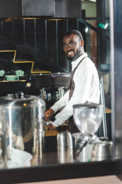 istock handsome young african american barista in eyeglasses smiling at camera while making coffee at coffee machine 961982138