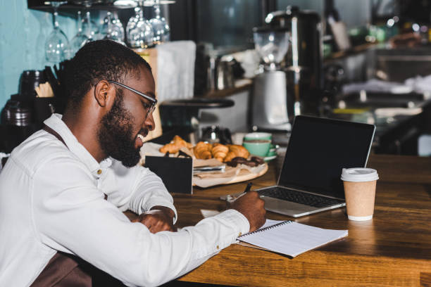 istock smiling young african american barista in eyeglasses taking notes and using laptop with blank screen in cafe 961983774
