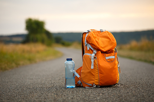 a backpack for an orange camera and a bottle of water on an asphalt road in a field at sunset