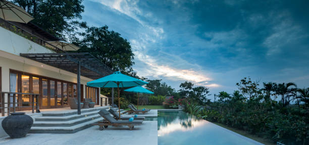 Located near Uluwatu in Bali, a luxurious villa with a swimming pool and lounge chairs is illuminated during a cloudy sunset. The real estate property faces south with a stunning view on the Indian ocean