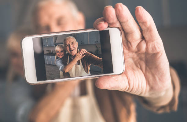 Joyful senior married couple photographing themselves on smartph Happy mature husband and wife are making selfie on mobile phone in kitchen. They are hugging and smiling. Focus on screen selfie photos stock pictures, royalty-free photos & images
