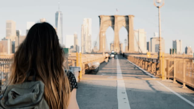 Camera follows young woman with backpack walking along Brooklyn Bridge, New York on a beautiful sunny summer day 4K