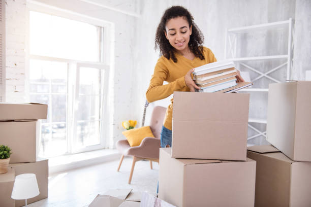Cute curly-haired girl packing books before moving out Favorite books. Pleasant curly-haired girl packing a pile of books into a big box before moving out of the room in the dormitory college dorm photos stock pictures, royalty-free photos & images