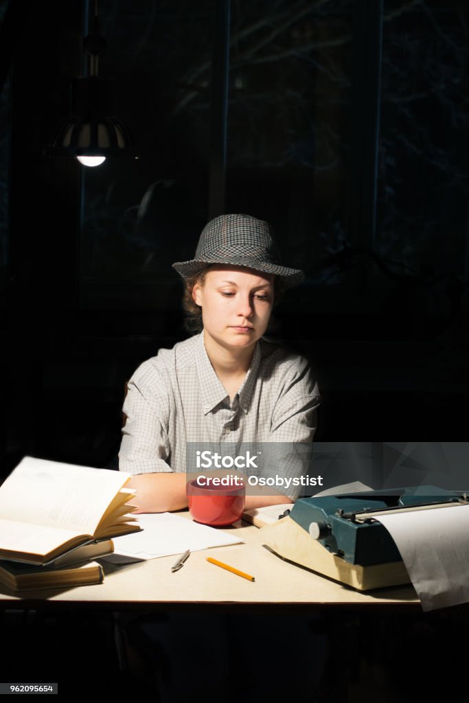 Portrait of a girl sitting at a table with a typewriter and books, think about the idea at night Portrait of a girl in a hat sitting at a table with a typewriter and books, think about the idea at night Adult Stock Photo