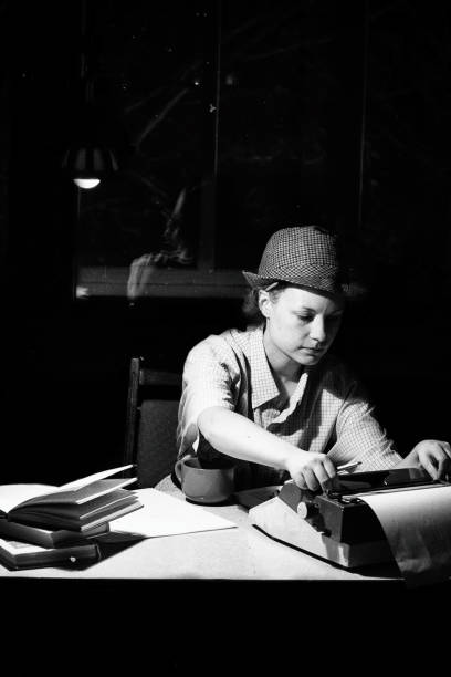 ritratto di una ragazza con un cappello seduto a un tavolo e digitando su una macchina da scrivere di notte - author writing retro revival women foto e immagini stock