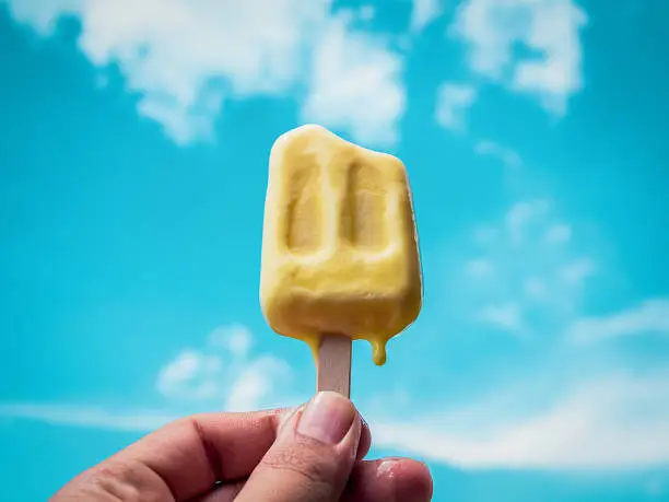 Hand Holding Yellow Frozen Popsicle Ice Pop Melting on Summer Blue Sky Background