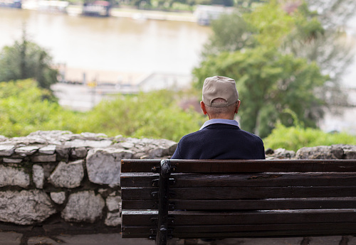 A lonely old man sitting on a bench in a park, looking at river