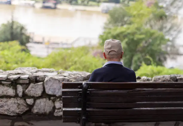 Photo of A lonely old man sitting on a bench in a park, looking at river