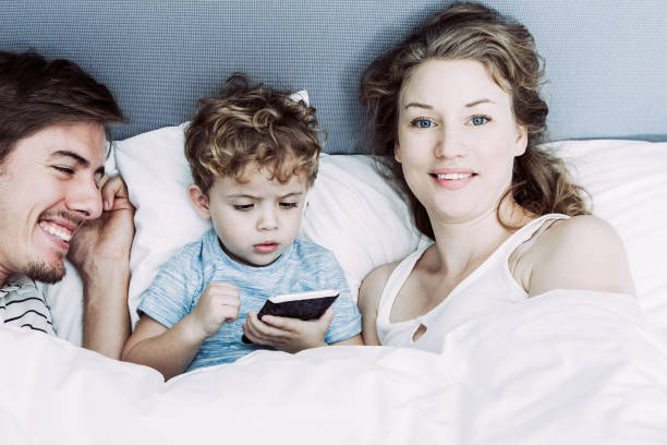 istock Funny boy holding phone lying in bed with parents 962146806