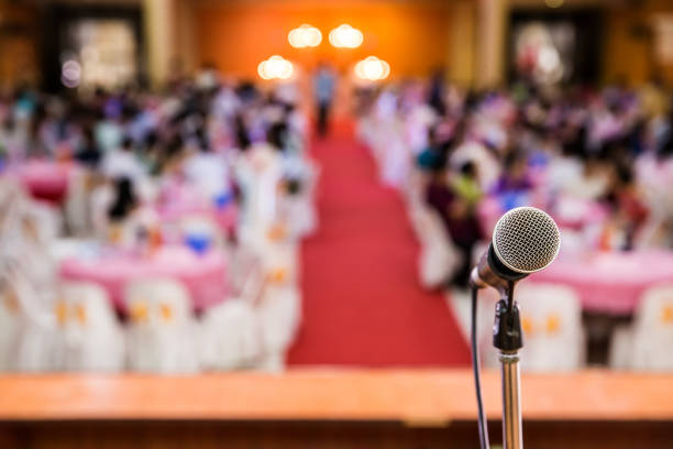 A close-up of a microphone on a podium, facing towards an audience seated in red chairs within an auditorium, ready for an event or presentation, with a blurred background.