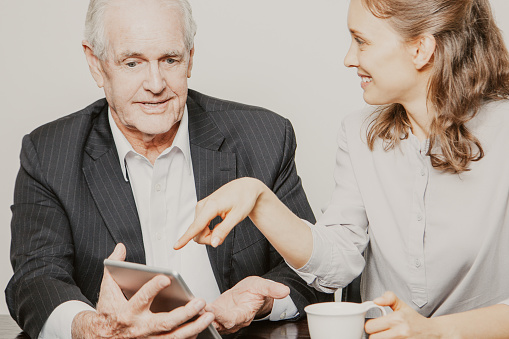 Closeup of smiling senior business man and female colleague showing something on touchpad screen. They are sitting at table and discussing ideas. Isolated view on white background.