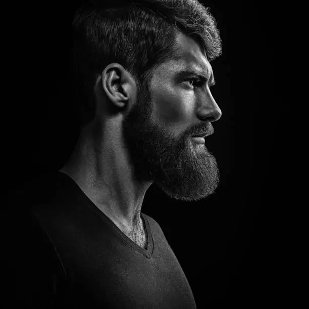 Dramatic concept Black and white close-up portrait of young handsome bearded man looking forward. Studio shot on black background