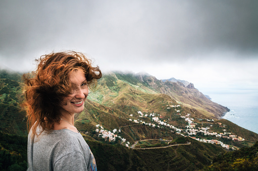 Joyful young woman with flying hair hidden her face looks at the camera in front of Taganana village on north of Tenerife. Concept of travelling, happiness, freedom. Traveler are enjoying the landscape, Canary Islands, Spain