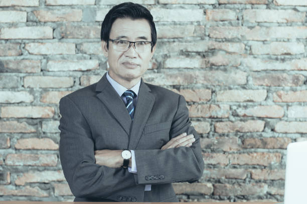 Serious senior businessman in modern office Portrait of serious senior Asian businessman wearing glasses sitting with folded arms at his workplace in modern loft office and looking at camera. Laptop and paper lying on table vietnamese old man portrait stock pictures, royalty-free photos & images