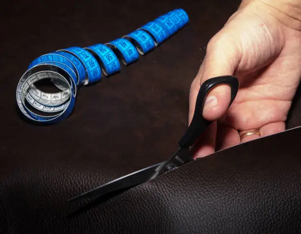 Cutting-out of leather using blue twisted tape measure and black scissors. Closeup