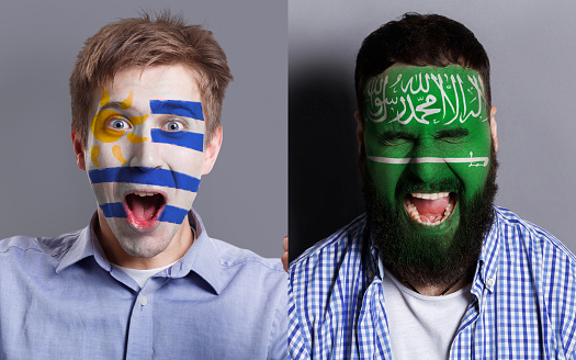 Emotional soccer fans with painted Uruguay and Saudi Arabia flags on faces. Confrontation of football team supporters from rival countries, sport event, faceart and patriotism concept.