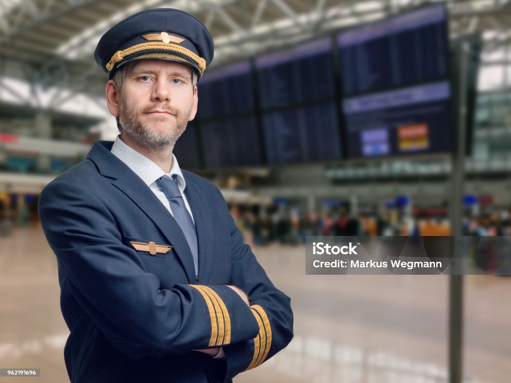 Pilot in uniform with golden stripes and cap crossed his arms while standing in the airport Portrait of a pilot in uniform with golden stripes and cap crossed his arms. It is located in the terminal of an airport Pilot Stock Photo