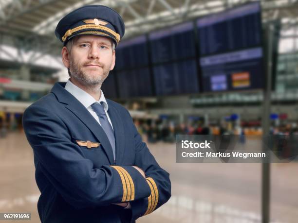 Pilot In Uniform With Golden Stripes And Cap Crossed His Arms While Standing In The Airport Stock Photo - Download Image Now