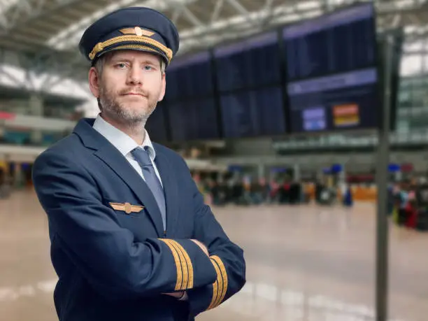 Photo of Pilot in uniform with golden stripes and cap crossed his arms while standing in the airport