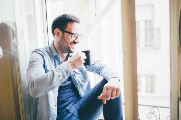 istock Happy young man with coffee mug sitting by window 962197124