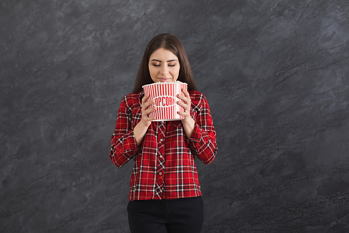 Happy beautiful young girl smelling popcorn with closed eyes, while watching film on grey background
