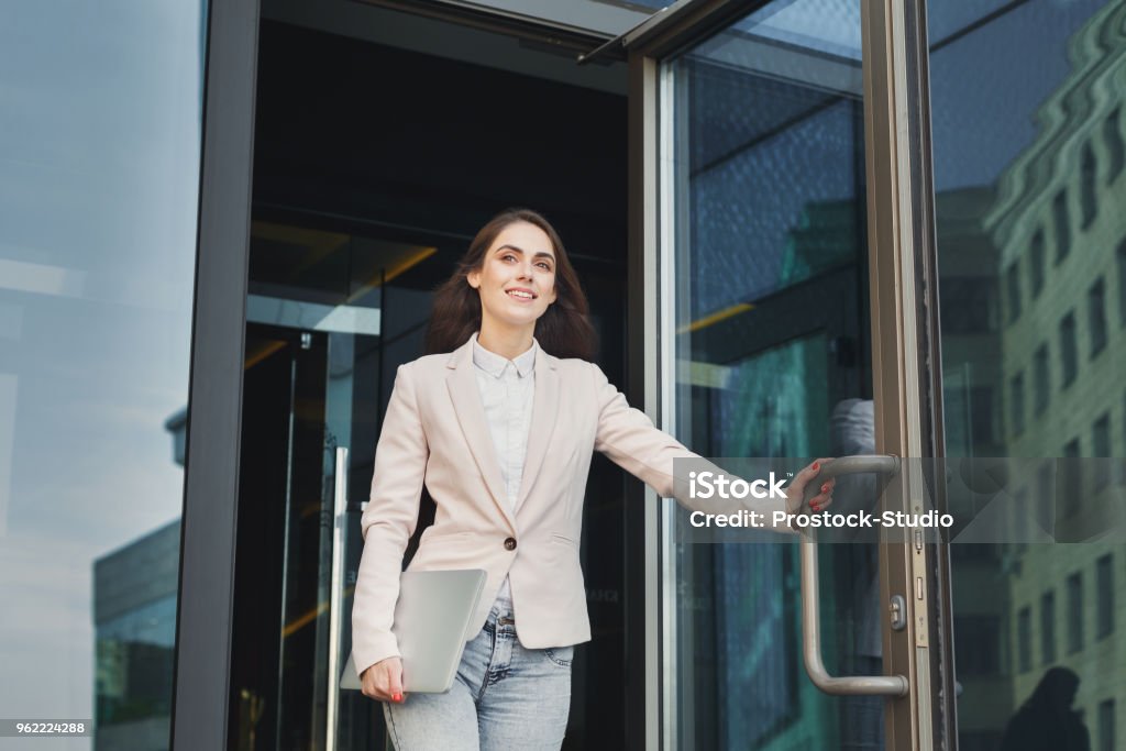 Confident young businesswoman talking on mobile Happy confident businesswoman walking out of modern office center, holding laptop outdoors, copy space Leaving Stock Photo