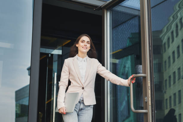 Confident young businesswoman talking on mobile Happy confident businesswoman walking out of modern office center, holding laptop outdoors, copy space goodbye stock pictures, royalty-free photos & images