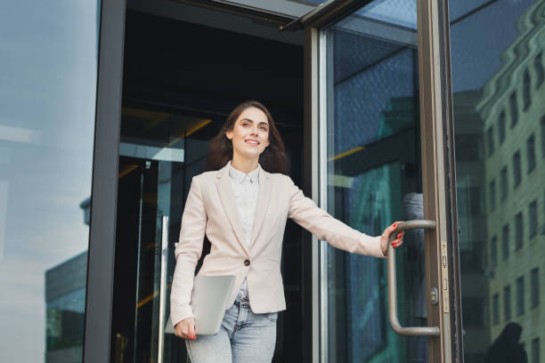 Confident young businesswoman talking on mobile Happy confident businesswoman walking out of modern office center, holding laptop outdoors, copy space office leave stock pictures, royalty-free photos & images