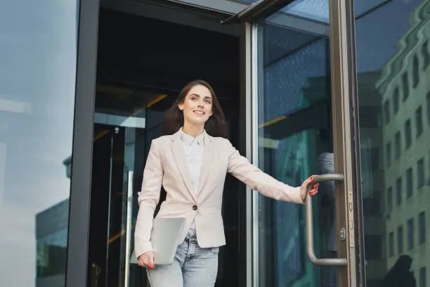 Happy confident businesswoman walking out of modern office center, holding laptop outdoors, copy space
