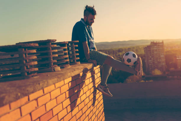 Football player holding a ball Football player on a building rooftop, sitting and relaxing after the match, holding a ball quiete socccer player stock pictures, royalty-free photos & images
