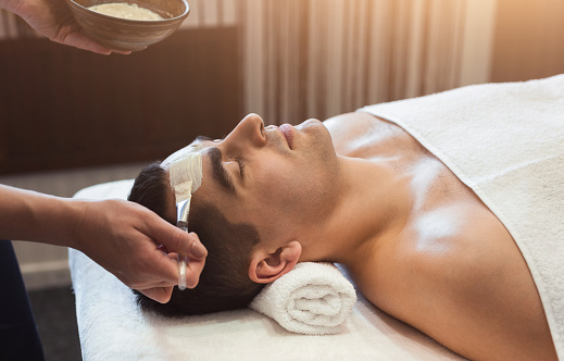 Man getting facial nourishing mask by beautician at spa salon, closeup. Apply face mask, spa beauty treatment and skincare.