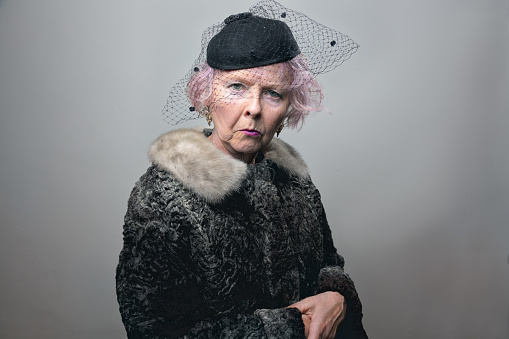 senior woman, wearing fur coat and black hat with veil, with pink hair, standing with a gesture of loneliness, looking with a serious or sad expression