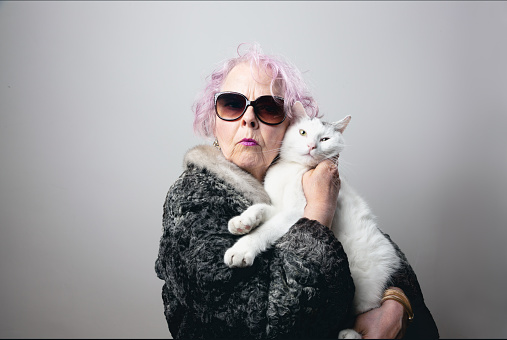 Senior woman, wearing olf fashioned fur coat and pink hair, holding white cat with black spots