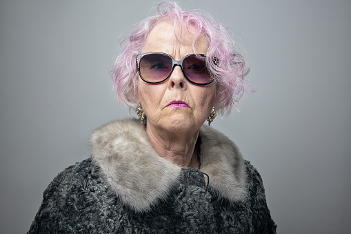 Senior woman with pink hair wearing old fashioned fur coat and sunglasses, looking at camera with arogant expression