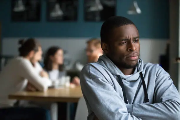 Frustrated excluded outstand african american man suffers from bullying or racial discrimination having no friends sitting alone in cafe, sad depressed black guy upset being rejected by white people