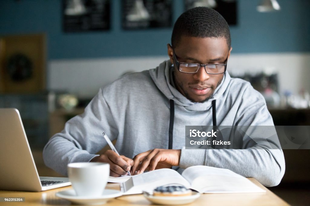 Focused millennial african student making notes while studying in cafe Focused millennial african american student in glasses making notes writing down information from book in cafe preparing for test or exam, young serious black man studying or working in coffee house Studying Stock Photo