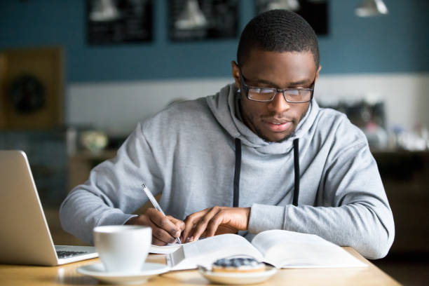 Focused millennial african student making notes while studying in cafe Focused millennial african american student in glasses making notes writing down information from book in cafe preparing for test or exam, young serious black man studying or working in coffee house exam student university writing stock pictures, royalty-free photos & images