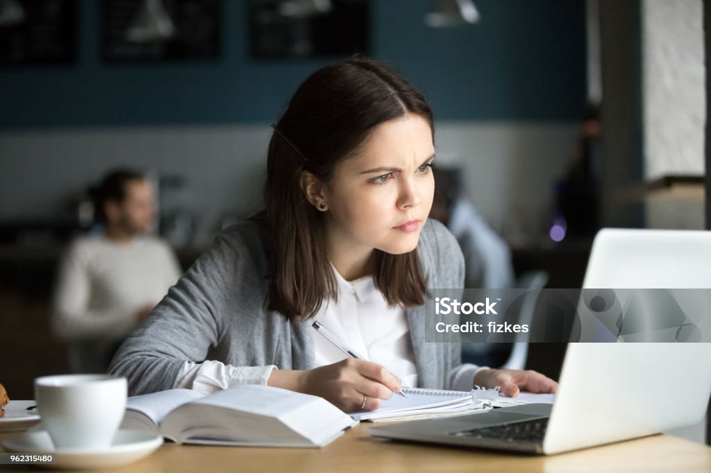 Focused girl concerned about difficult online task studying in cafe Focused girl concerned about difficult online assignment looking at laptop screen studying working in cafe, millennial student thinking of problem solution reading hard task on computer making notes Toughness Stock Photo