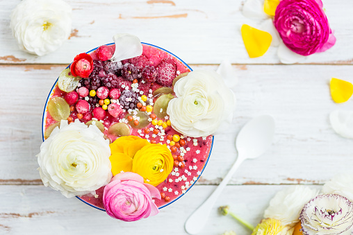 Breakfast Detox Pink Smoothie from Banana and Forest Berries in the Bowl, topped with frozen berries and ranunculus flowers, light wooden background, top view
