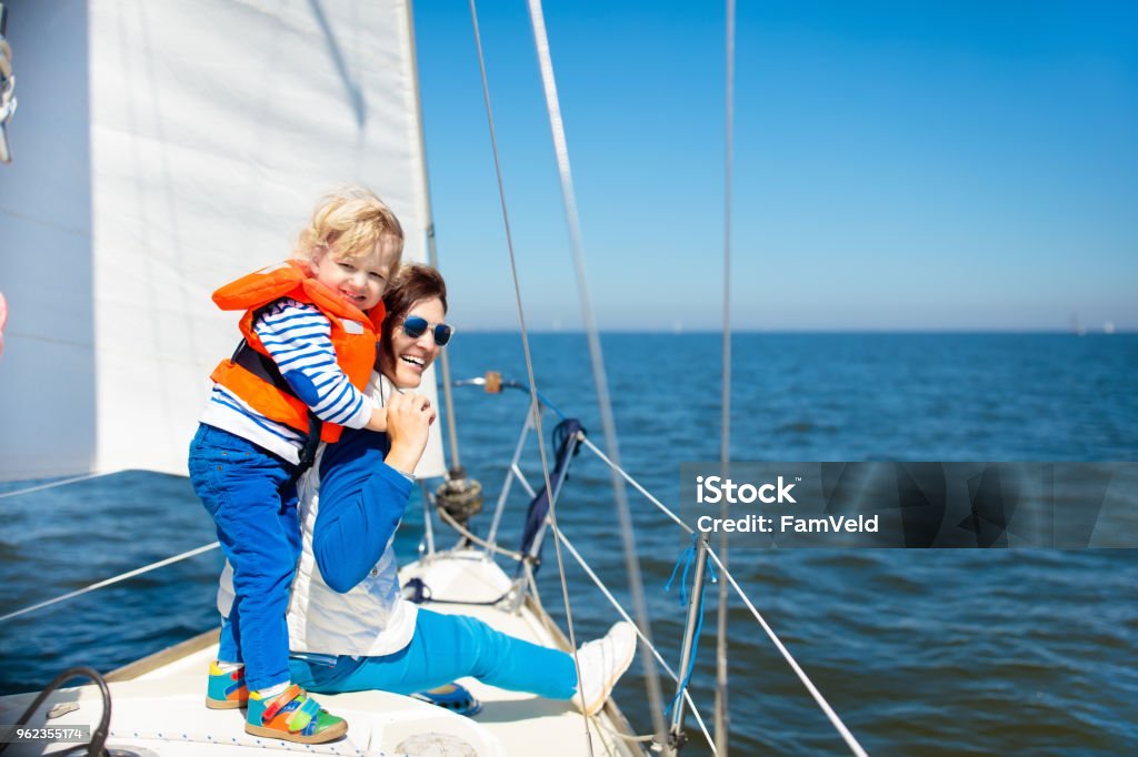 Family sailing. Mother and child on sea sail yacht. Mother and baby boy sail on yacht in sea. Family sailing on boat. Mom and kid in safe life jacket travel on ocean ship. Parent and child enjoy yachting cruise. Summer vacation. Sailor on sailboat. Nautical Vessel Stock Photo