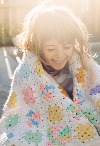 Sweet little 4 year old girl in early morning light outdoors and wrapped in a vintage crocheted blanket, perhaps handmade by a grandparent. She is a cheerful child