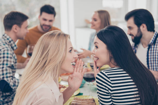 duas garotas atraentes, elegantes, melhores, segurando a palma perto do rosto sussurrando fofocas femininas uns aos outros sobre os seus amigos que sentado na mesa de jantar desfrutando, ceia - whispering couple discussion smiling - fotografias e filmes do acervo